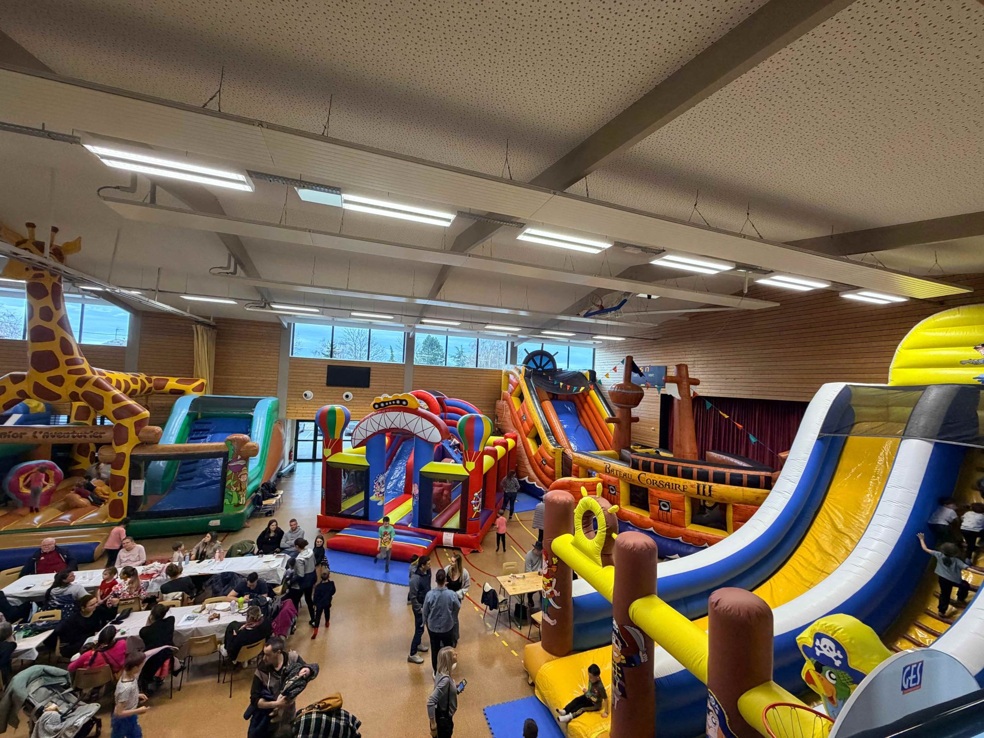 Vue d'ensemble du parc gonflable dans la salle polyvalente, avec la grande girafe, le Corsaire III, des toboggans et des familles